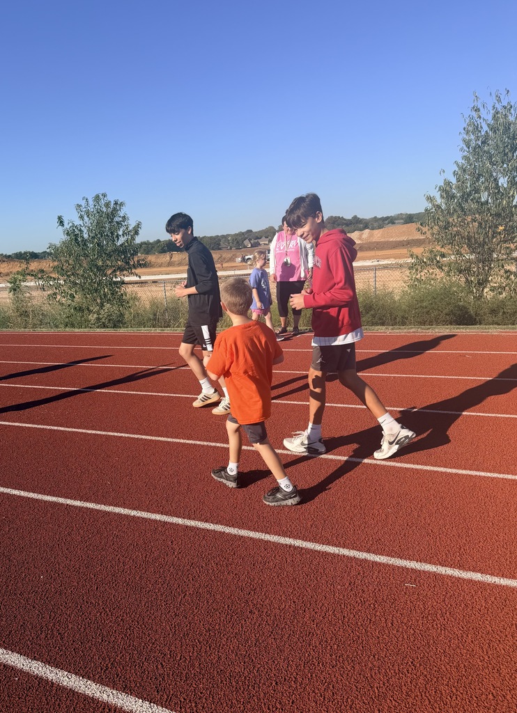 Several people, including a child, walk on a red track under a blue sky. Trees are in the background.