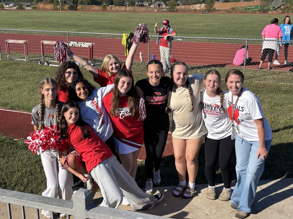 A group of individuals in red and white shirts pose for a photo near a metal fence. Behind them, a sports field and a grassy area are visible.