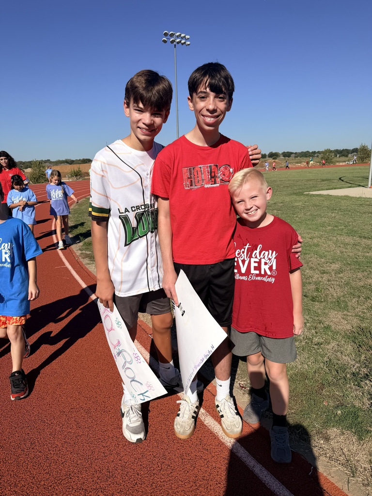 Three young boys stand together on a track, with two holding papers, one wearing a baseball jersey. A grassy field and track lights are behind them.