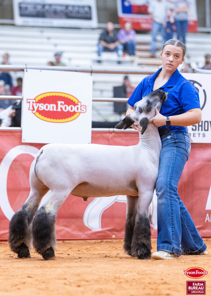 Woman with blue top and jeans holds lamb during an event with Tyson Foods banner in background.