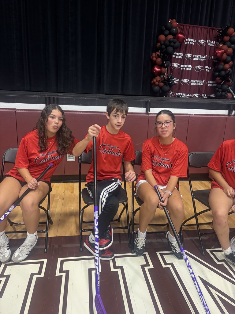 Four individuals in red shirts seated in chairs with hockey sticks, holding them, and a large sign in the background.