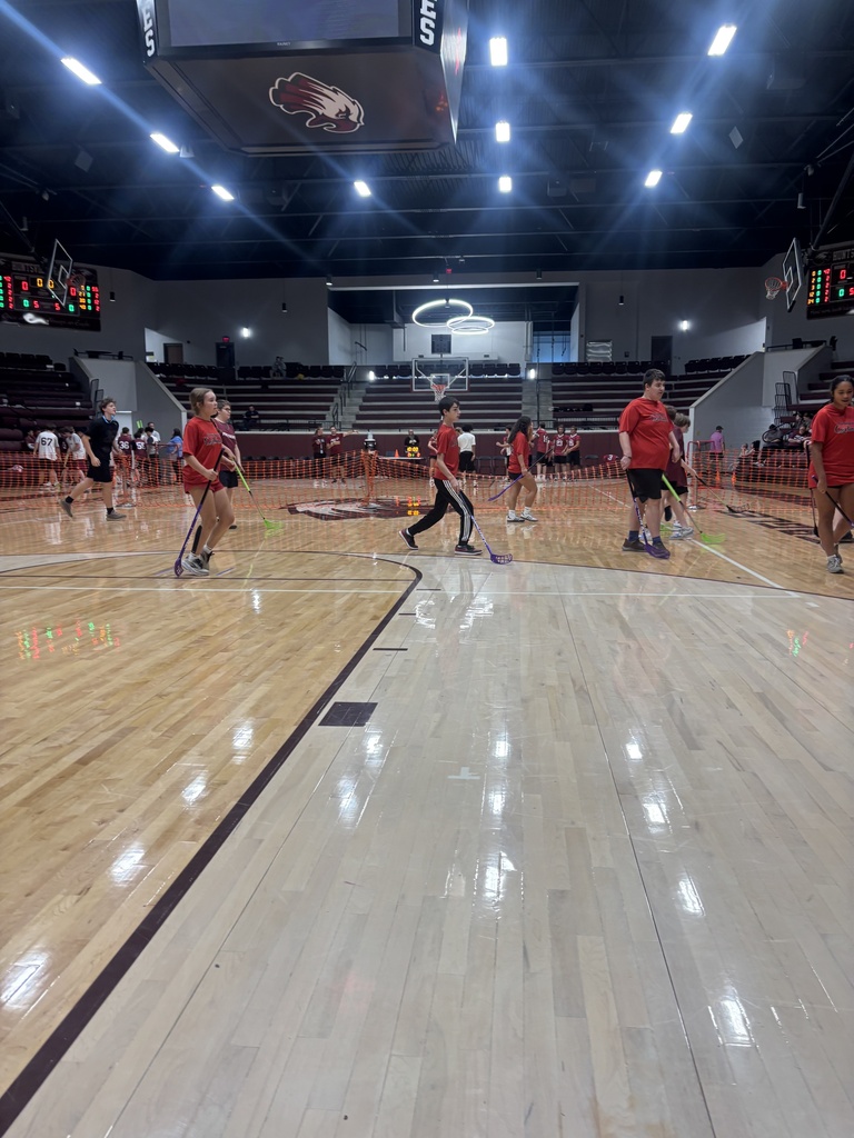 Several people wearing red shirts play broomball on a wooden floor in a gymnasium. The court has lines and some people are standing.