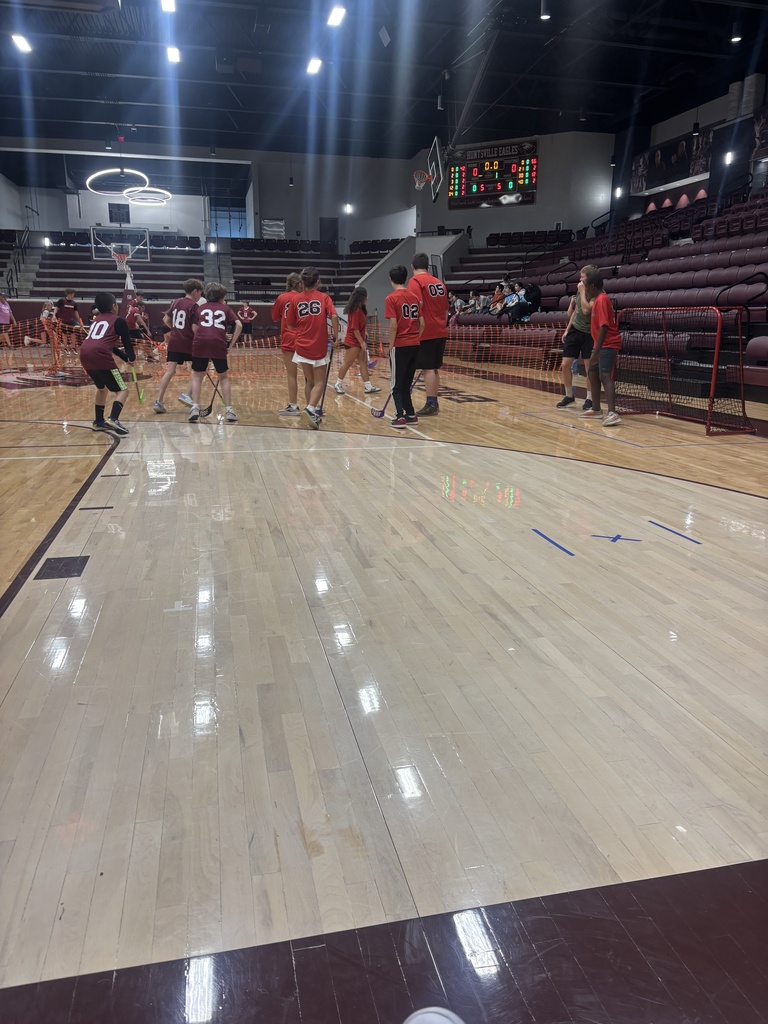 A basketball game in progress in a gymnasium with a hardwood court, players in red jerseys, and an empty seating area.