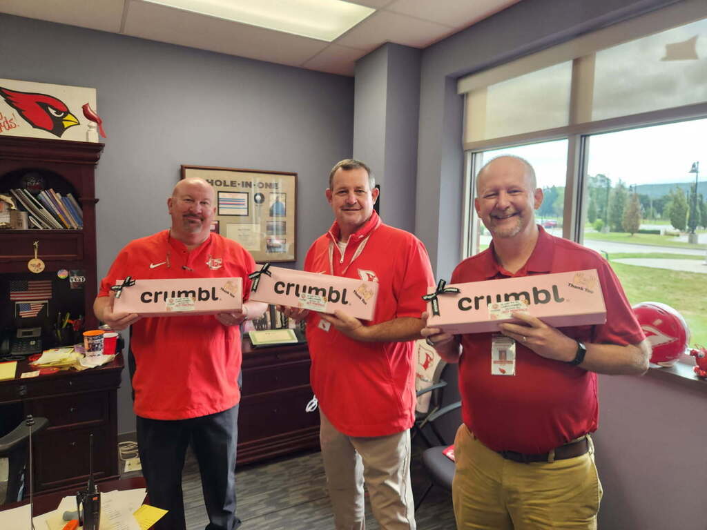 Three people in red shirts stand in an office with red boxes, reading "crumbl" in white letters. A window shows a view of trees.
