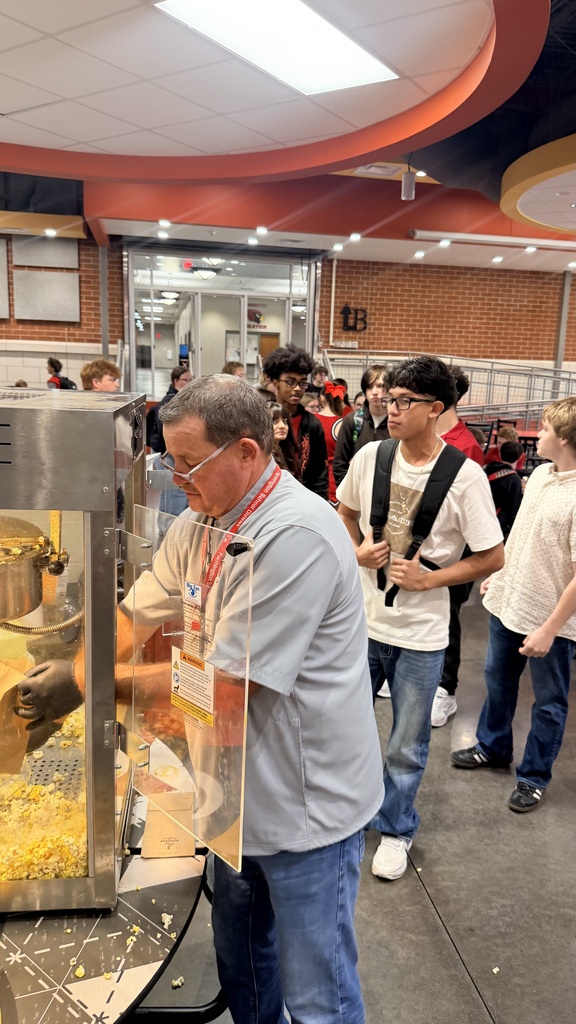 A man with glasses stands in front of a popcorn machine in a cafeteria while others wait.
