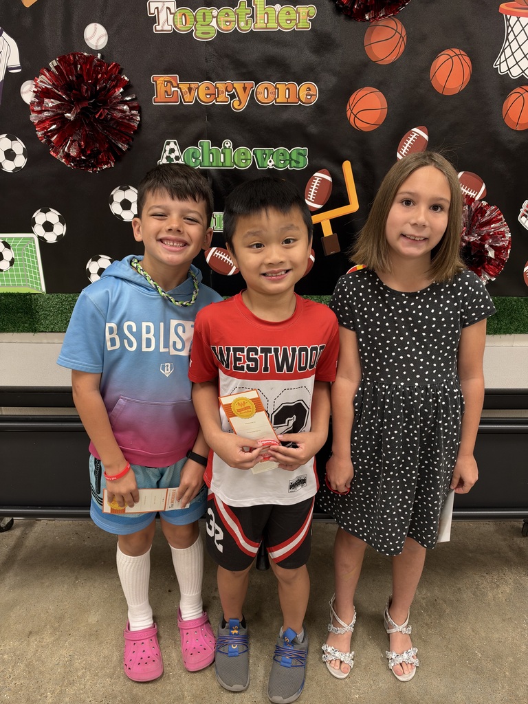 Three young kids stand close together in front of a sports-themed backdrop. Each holds a snack.