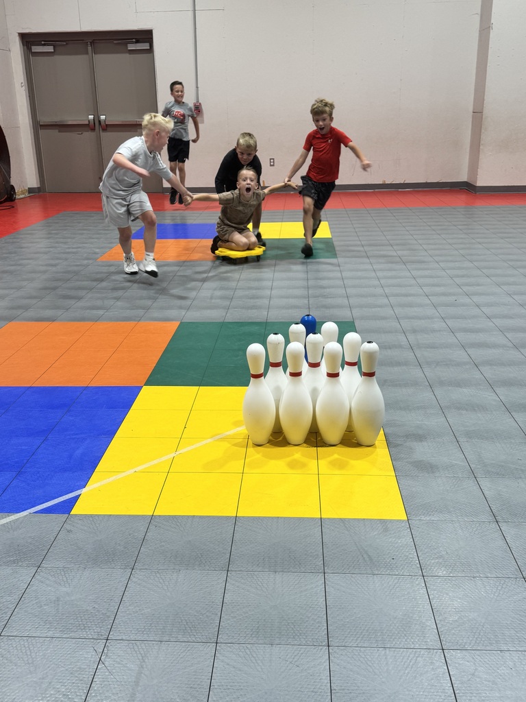 Four children playing on a multi-colored floor. One child sits on a scooter while another runs. Two adults supervise. Bowling pins are at the front.