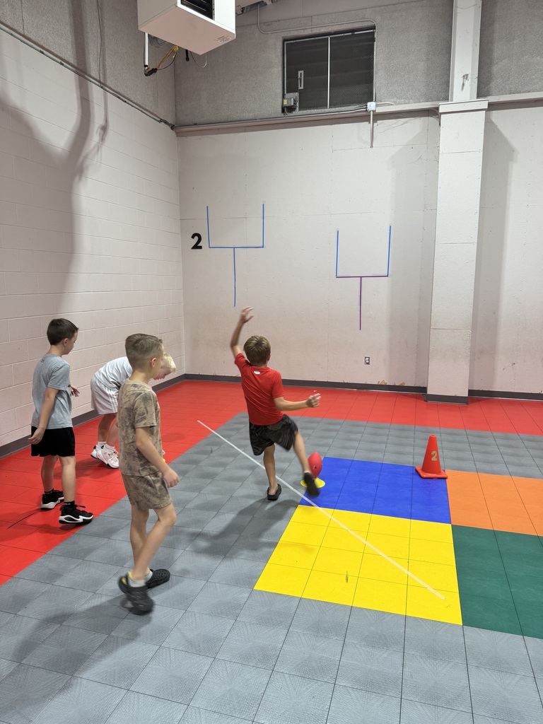 Four children play a game in a gym with white walls. A boy holds a ball and prepares to throw it. Multicolored tiles cover the floor.