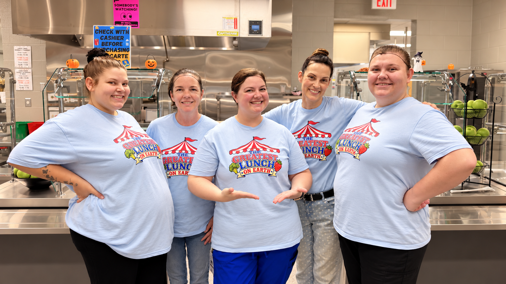 Five women in matching blue shirts stand in a kitchen, smiling. They wear black pants and blue pants.