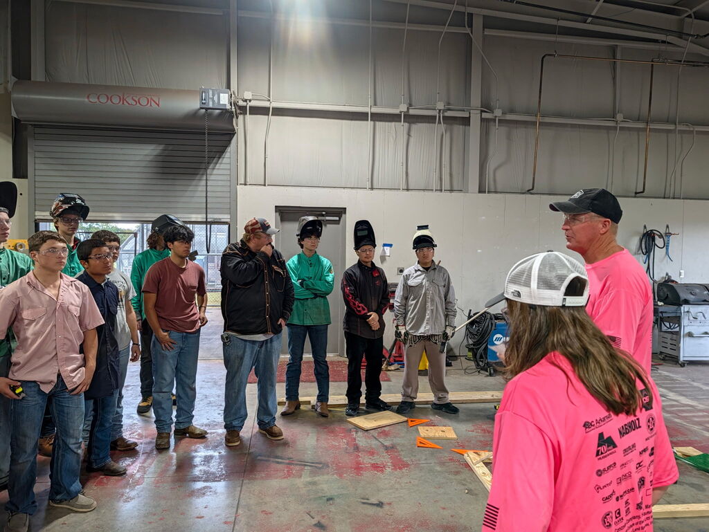 A group of people in a workshop, wearing pink shirts and hats, appear to be listening to a presentation.