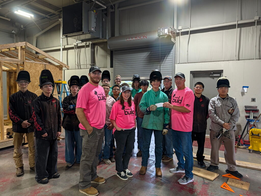 A group of people in pink shirts and helmets standing in a workshop. Behind them, a wooden structure is under construction.