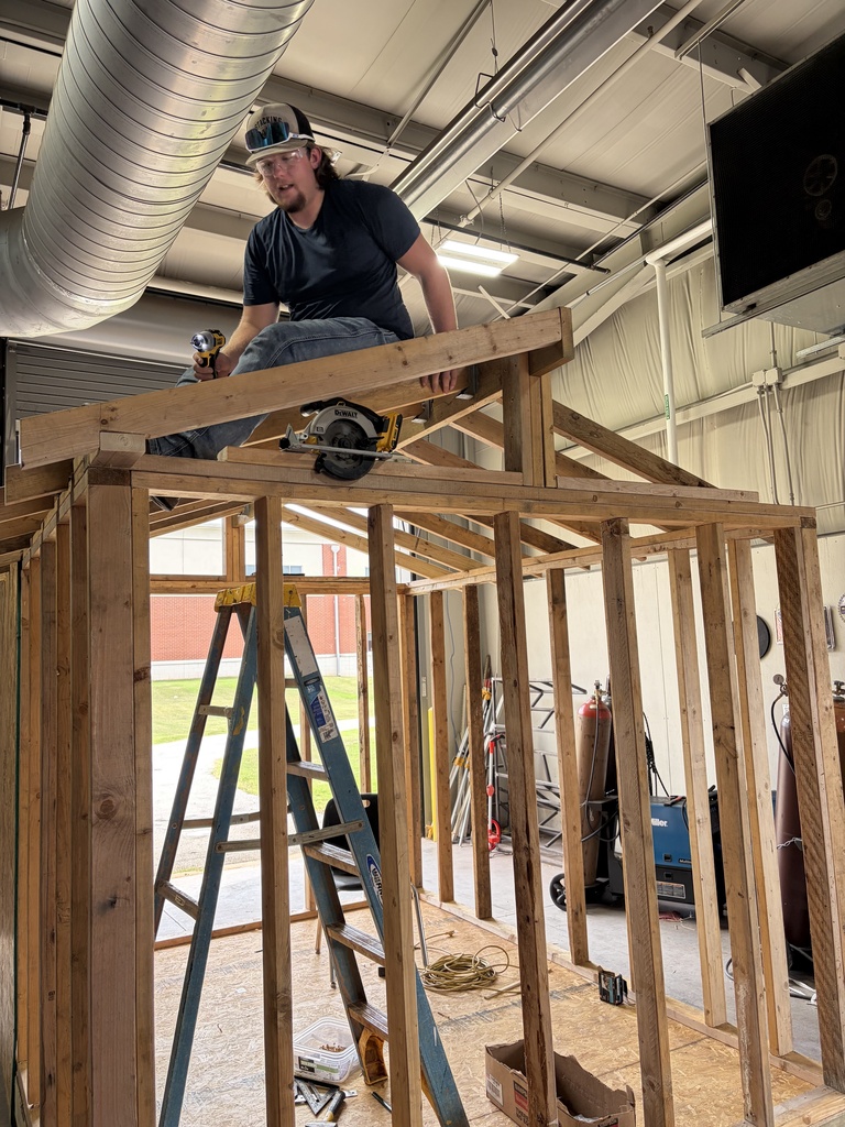 A man sits on a wooden structure under construction. He is wearing a helmet and safety goggles and holds a circular saw.