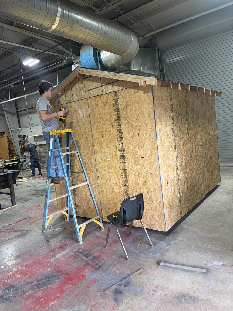A person on a ladder works on a wooden structure, likely a shed, in an industrial setting.