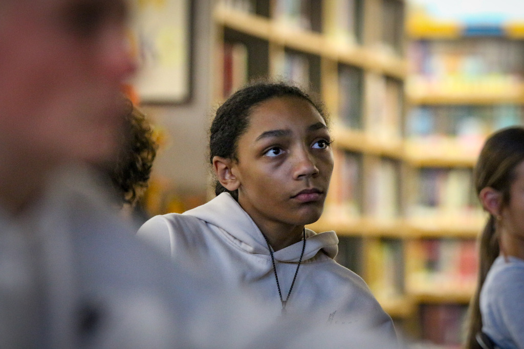 A student sits in a library facing forward, with several bookshelves behind them. Other students are partially visible.