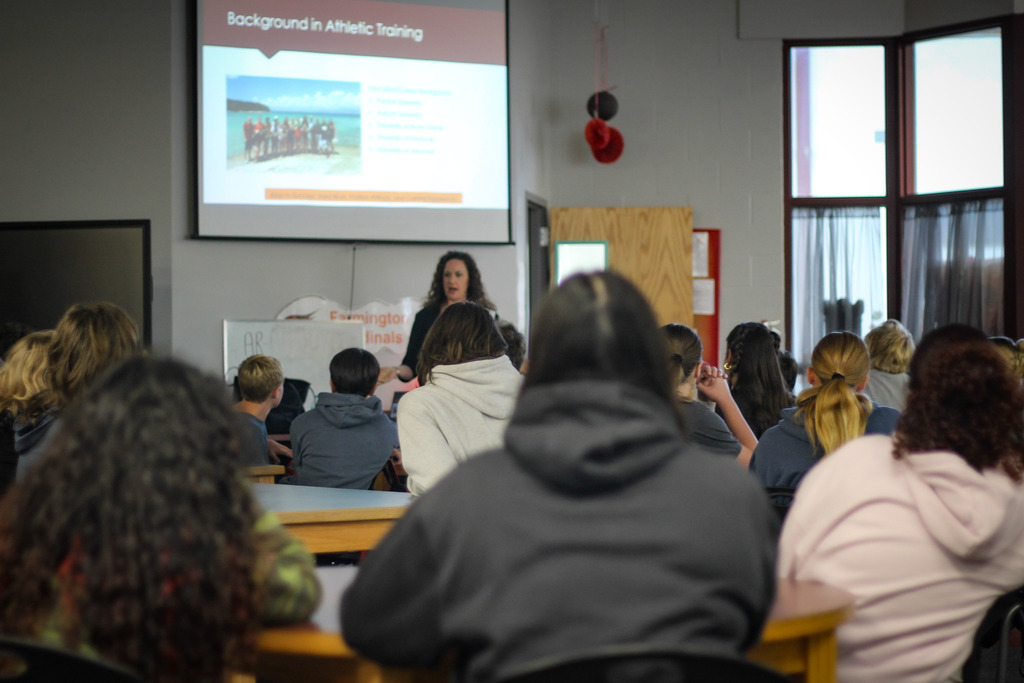 Woman standing in front of screen showing presentation, several students in chairs facing her, windows in background.