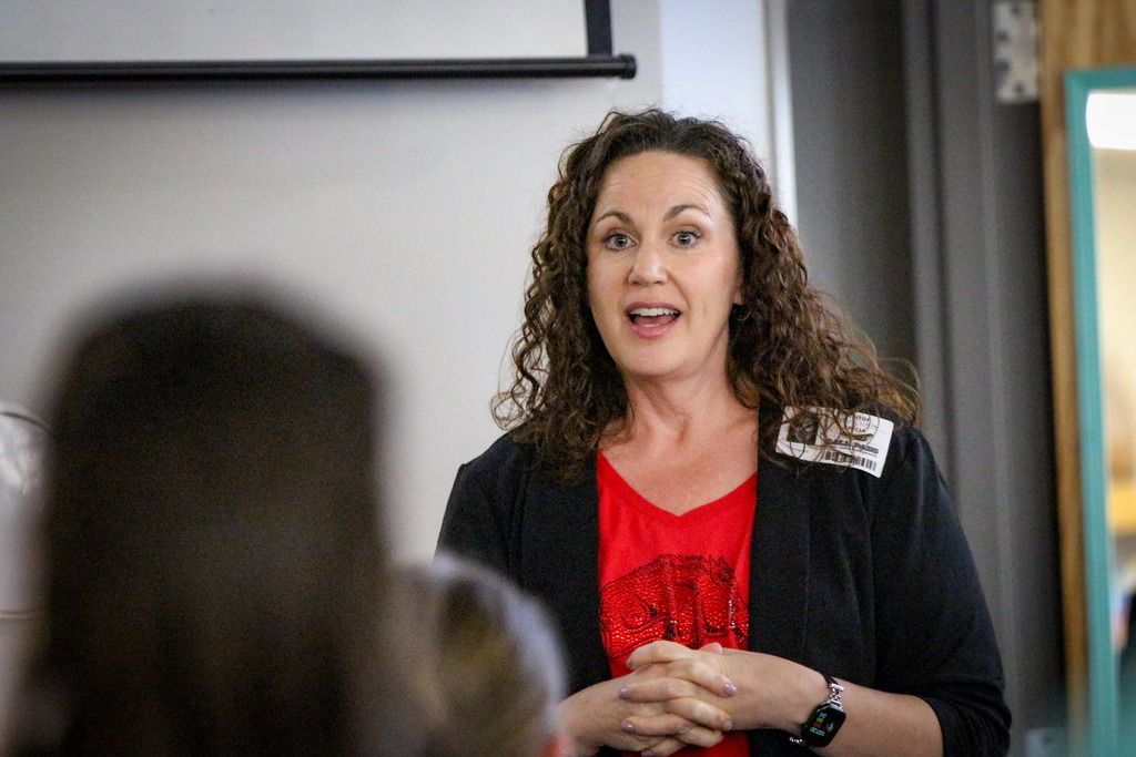 A woman with curly hair speaks to a group of people; she wears a red shirt and black cardigan.