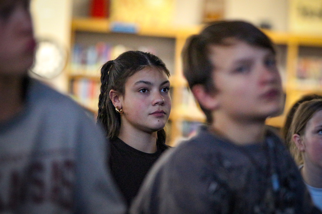 Four children facing forward, with a wooden bookshelf in the background with books visible on the shelves.