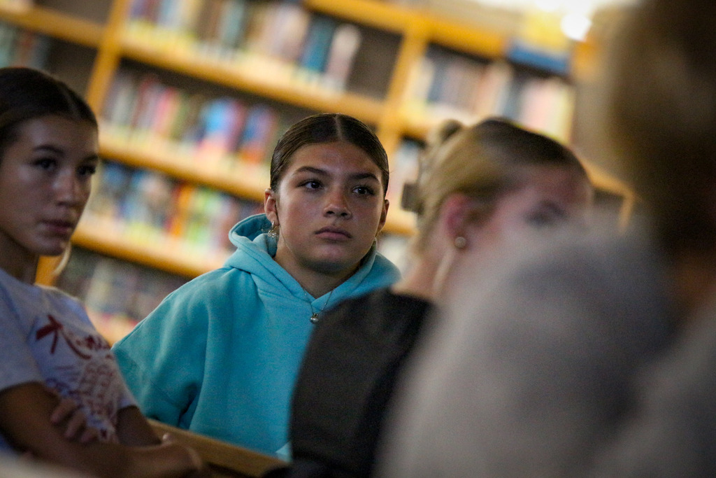 Four students face forward in a library. One in a blue hoodie looks serious. Behind them, a bookshelf filled with books.