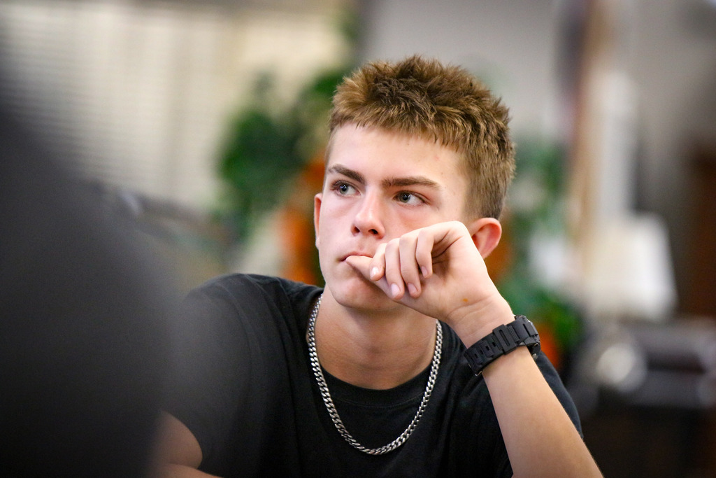 A young man wearing a black shirt and silver necklace sits with his chin resting on his hand.
