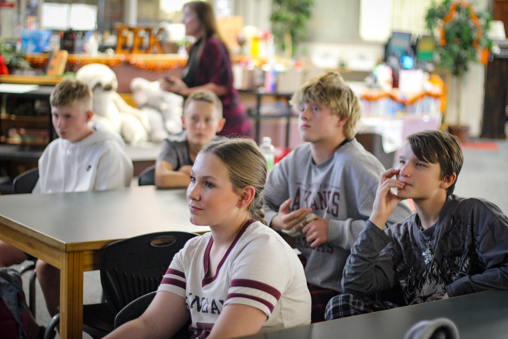 Group of children seated around tables, one boy with hands clasped on his chin, in a classroom with shelves and plants.