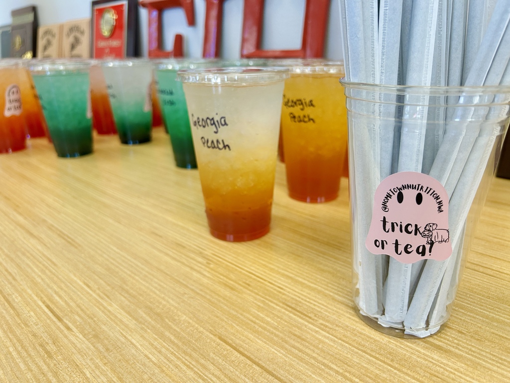 Multiple plastic cups with different colored beverages on a wooden table, a cup with "tricks or tea" label.