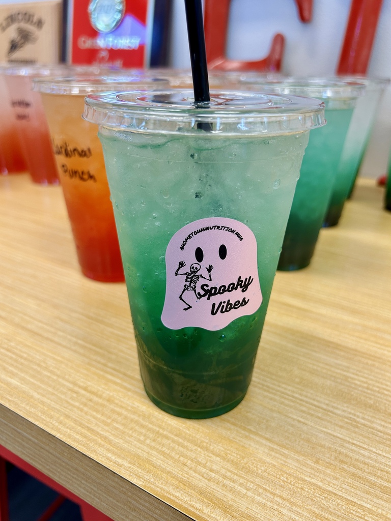 Four cups of drinks with straws sit on a table. The closest cup has green and blue liquid.