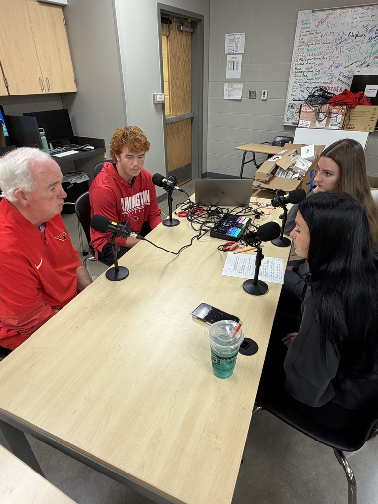 Four people sit around a table with microphones. Two have laptops, one holds a cup, and a whiteboard is in the background.