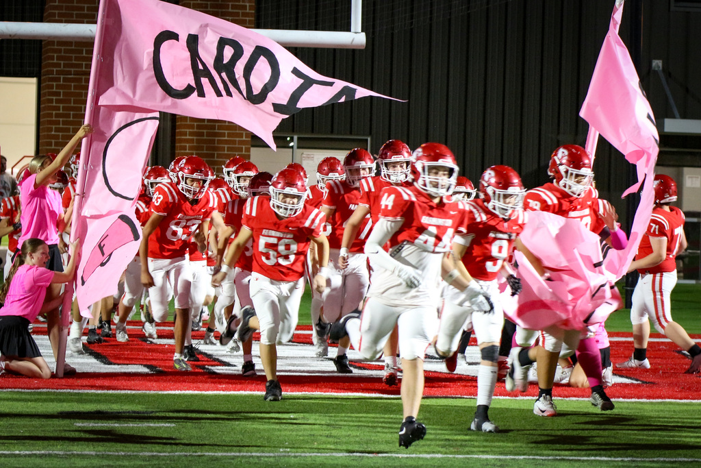 Football team with red helmets, jerseys, and white shorts running on a field, led by people waving pink flags.