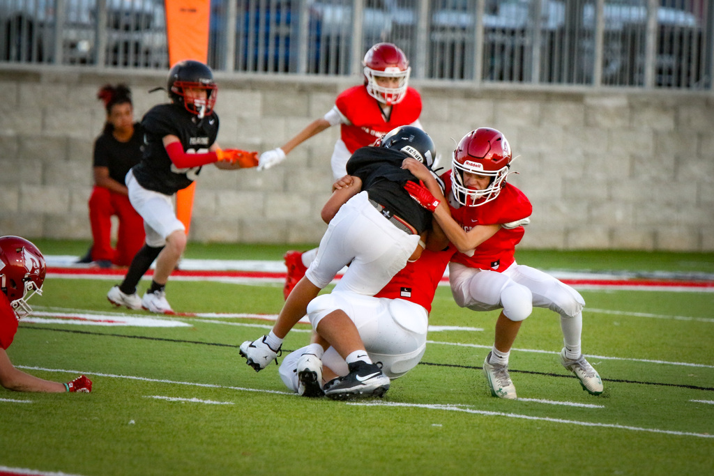 Youth football players wearing red and white uniforms, helmets, and gloves tackle and wrestle on the field.