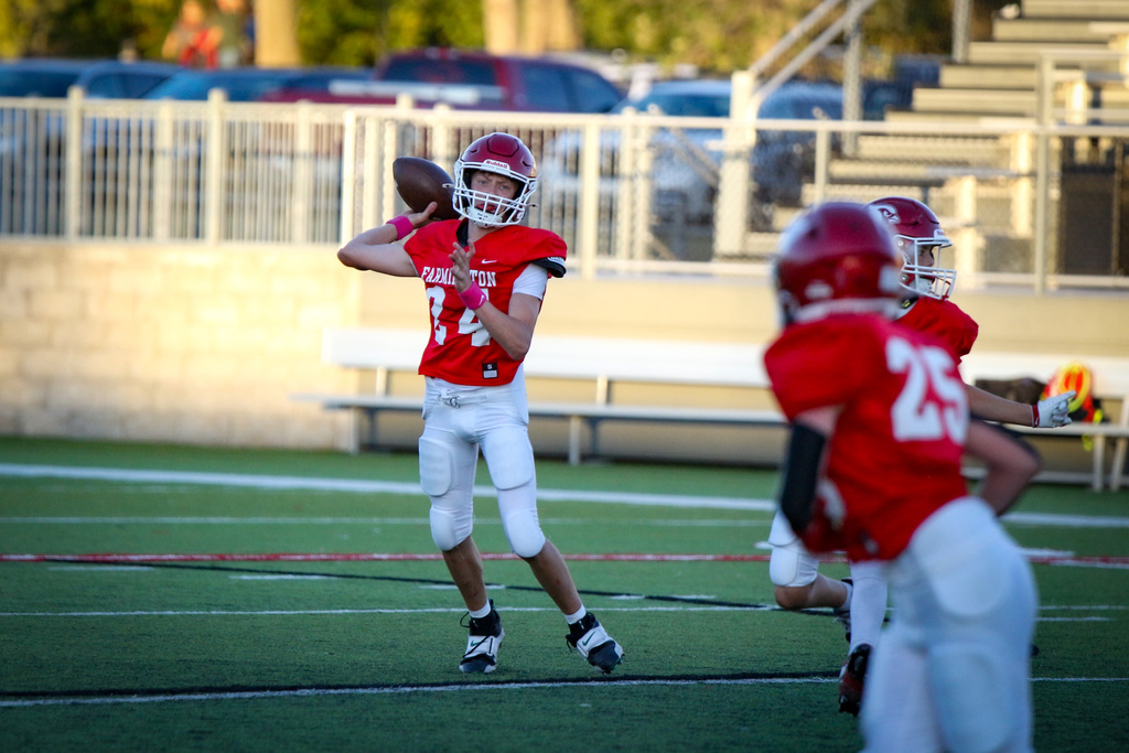 Football player in red uniform passes ball on a football field. Players in red helmets run in background.