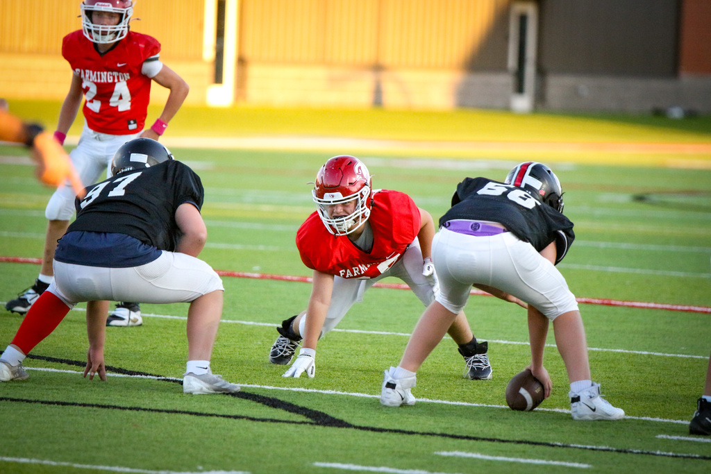 Five football players in uniforms lined up on field. One holds a football. Three have red helmets, two black.