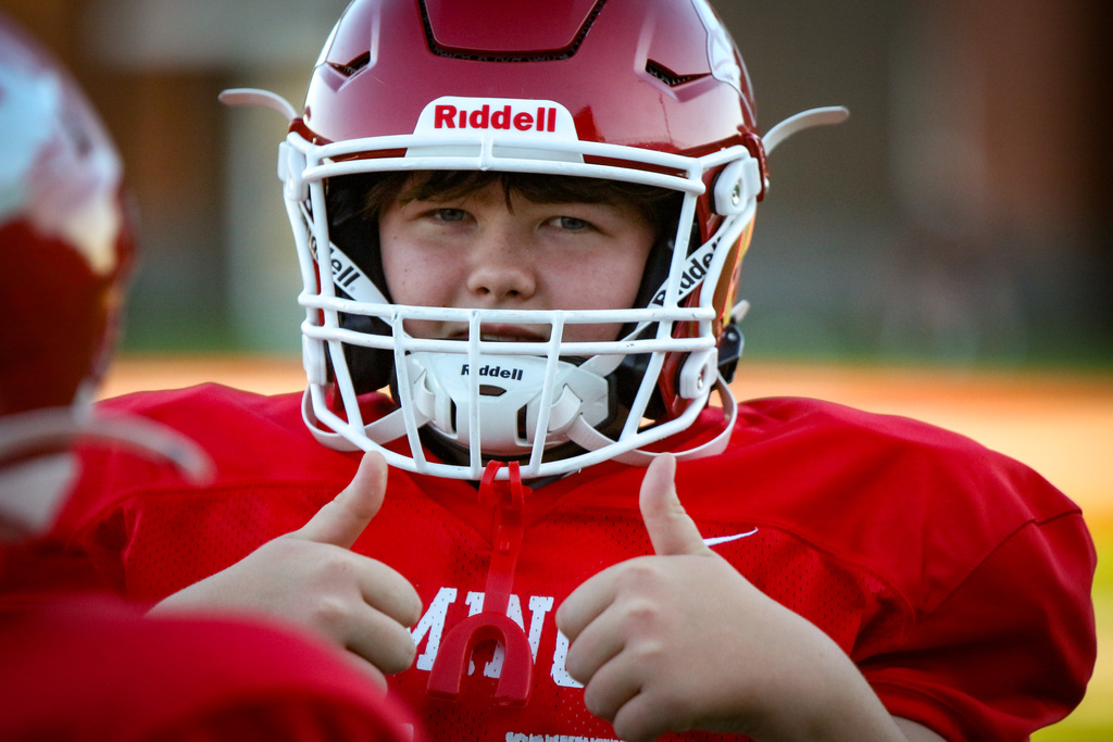 A youth football player in a red helmet and jersey giving two thumbs up, with another player partially visible.