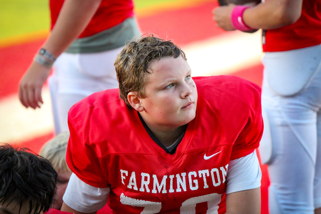 Young person in red jersey with word Farmington sitting on ground with others during game.