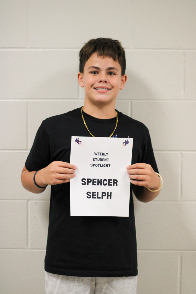 Boy in a black t-shirt holding a white sign with black letters reading "Weekly Student Spotlight Spencer Selph".