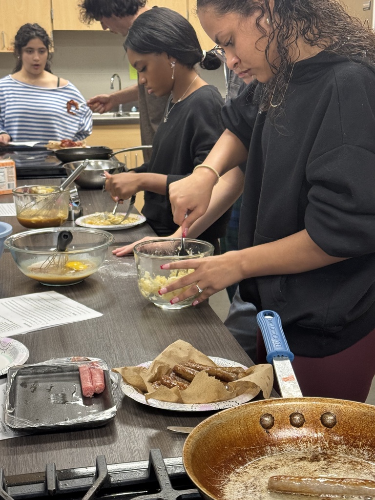 Several students work in a kitchen, cooking food on the stove and preparing ingredients at a table.