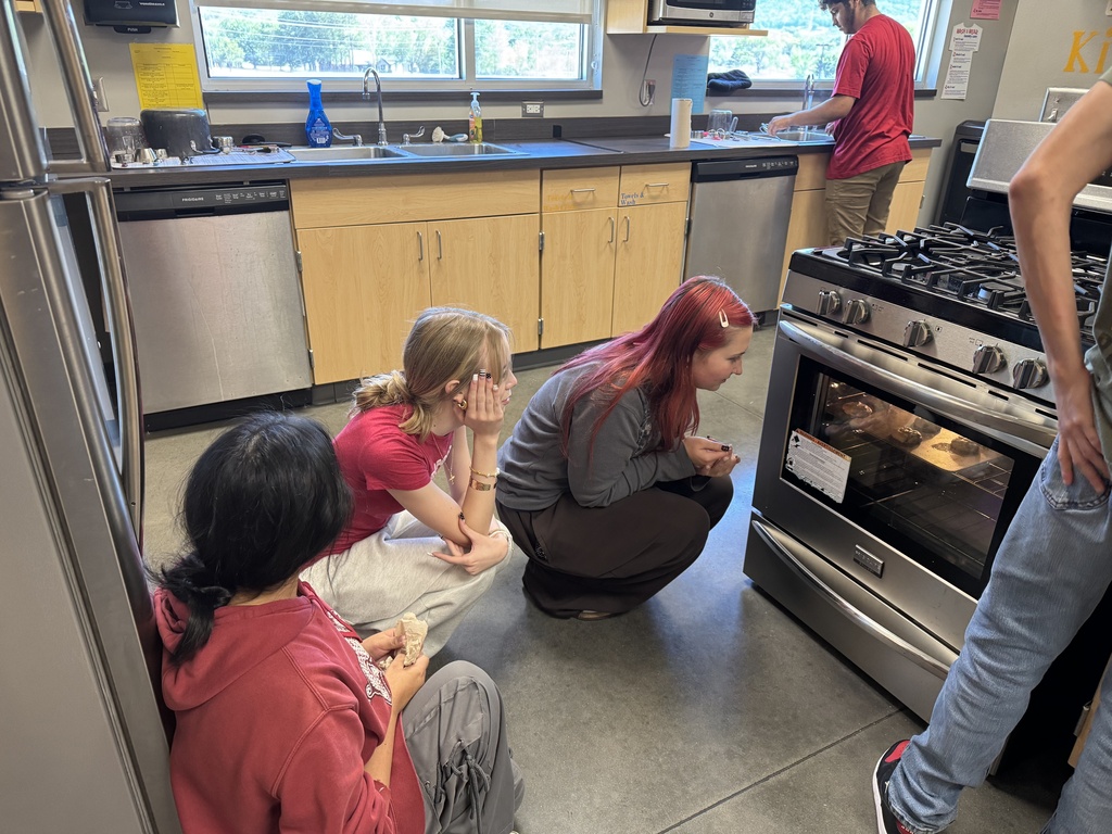 Four students in a kitchen, three sitting and one crouching. A gas stove with an oven door open.