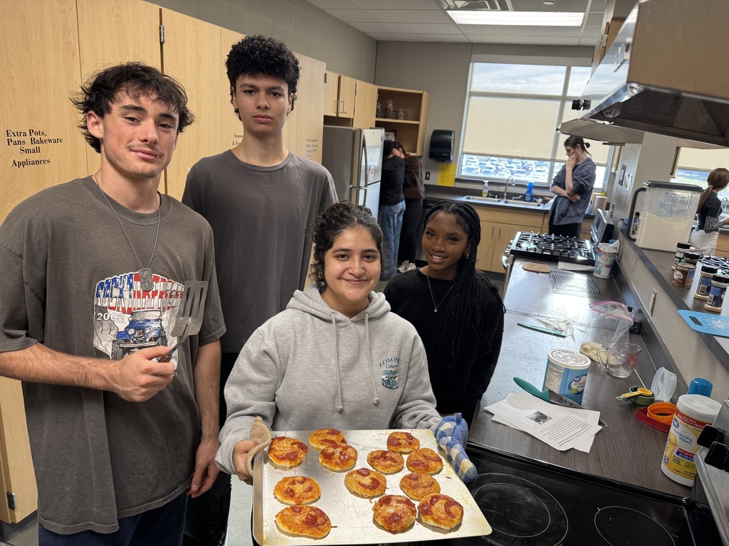 Four students stand in a kitchen. One holds a tray of golden-brown pastries. A sink, stove, and cabinets are in the background.