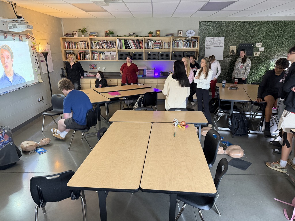 A classroom with tables and chairs, a projector screen, and people gathered around. One person sits on a chair.