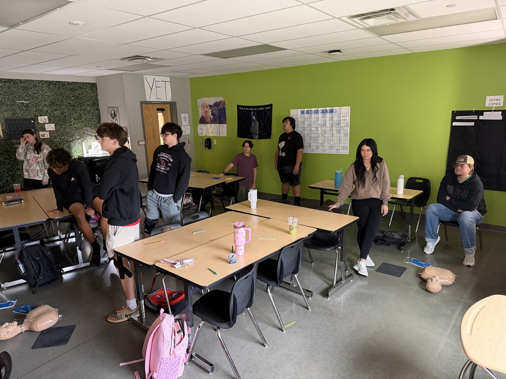 Students in a classroom with green walls. A group stands near tables, chairs, and backpacks.
