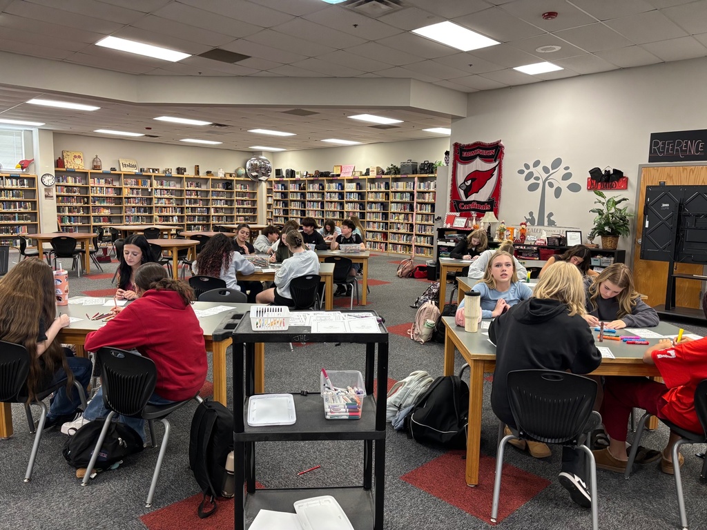 Students seated at tables in a library, some with bags, engaged in reading and writing.