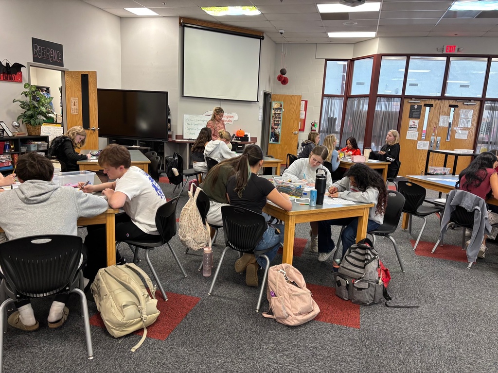 Students sit at tables in a classroom, writing with pens. Backpacks and bags are on the floor.