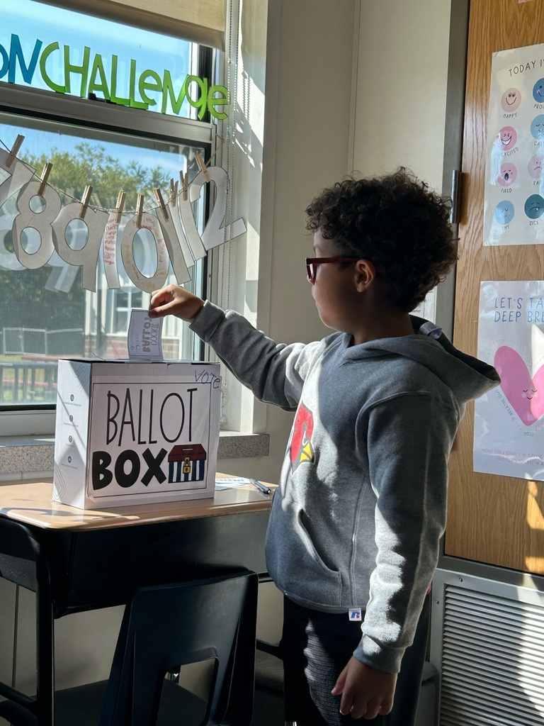 Child in hoodie places paper into a box labeled Ballot Boxmm. Box is on table in front of window.