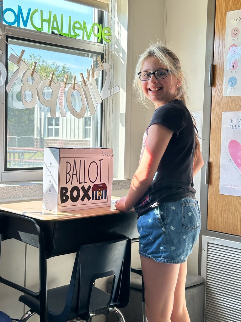 Girl standing next to a box labeled "BALLOT BOX" with a smile on her face.