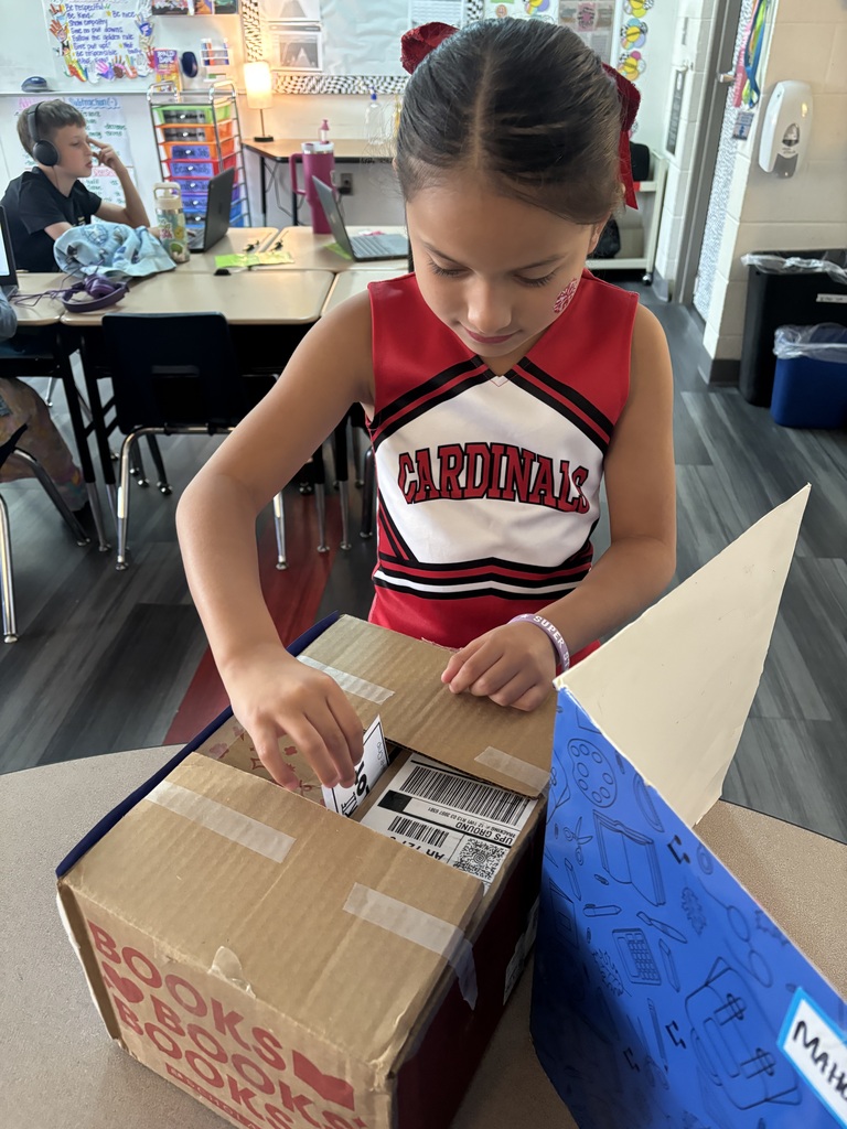 Girl in red and white cheerleading uniform opens a box labeled "Books" on a table.