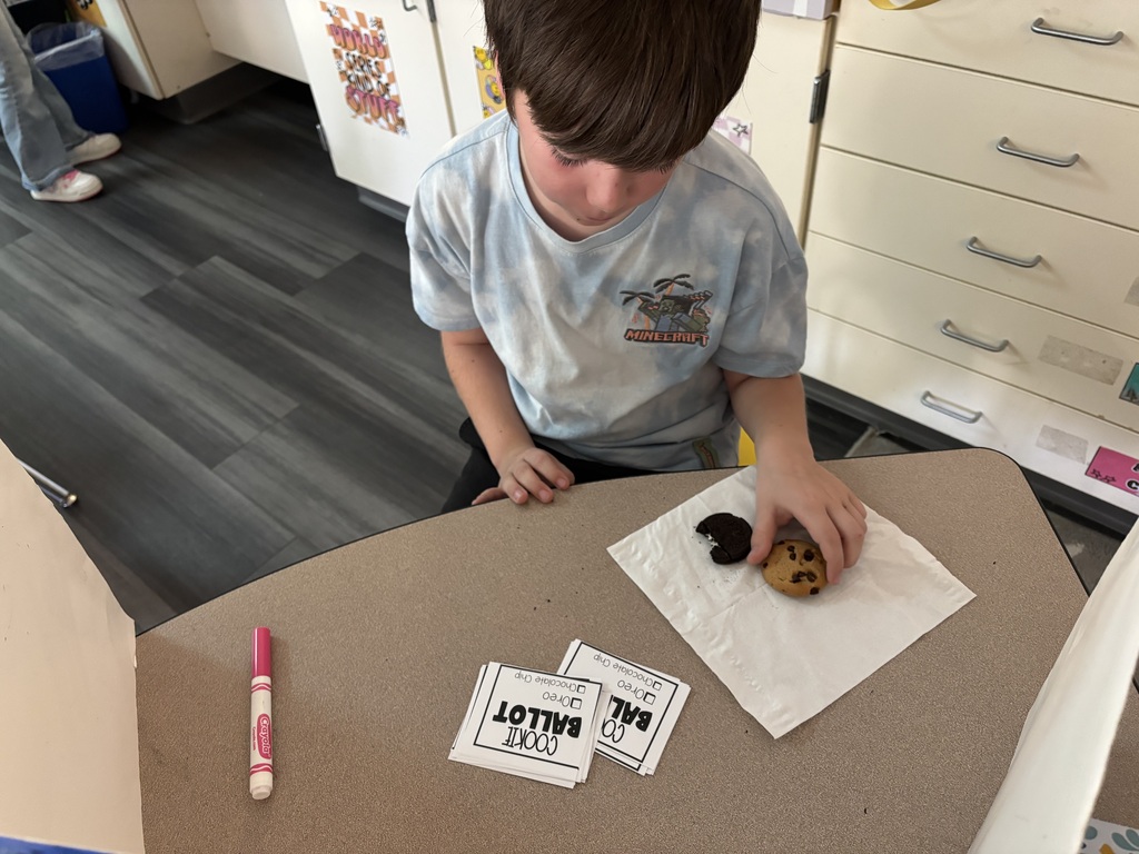 A young boy sits at a table and holds a cookie. A napkin, markers, and cards are on the table.