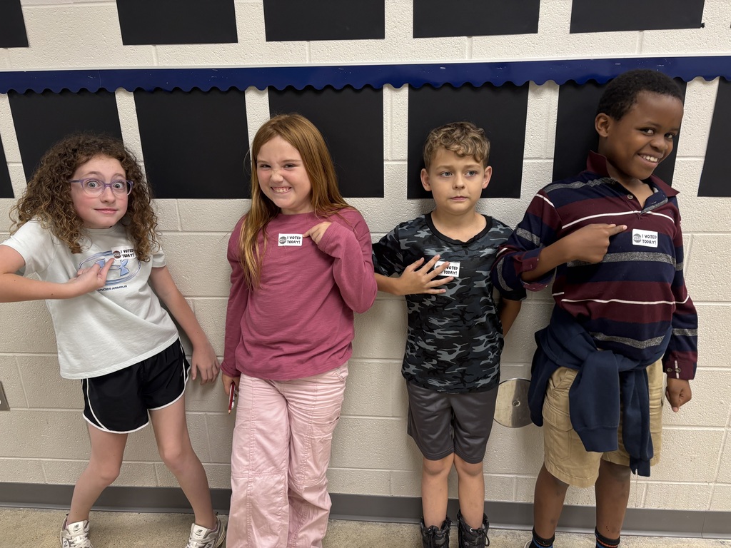 Four children stand next to each other against a white and blue wall. One points at something.