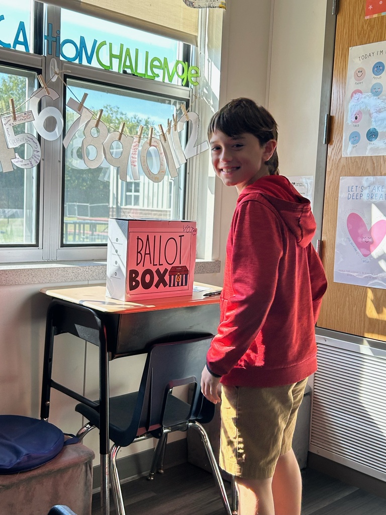 Boy in red hoodie standing by a desk with a ballot box on it.