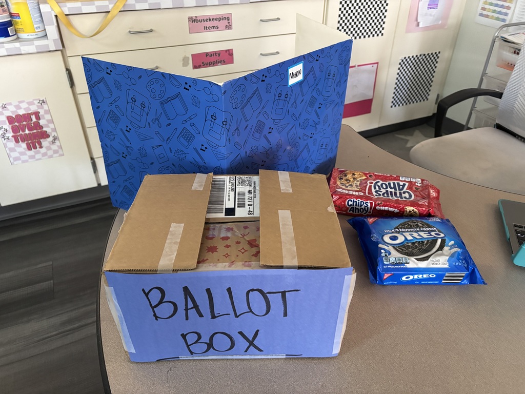 Cardboard box labeled "ballot box" with an open blue folder and cookies on a table in a classroom.