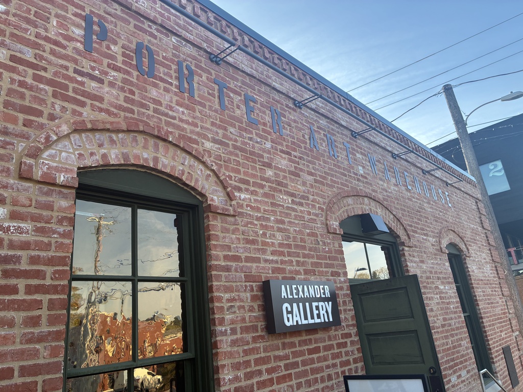 A brick building with arched window, arched doorway, and sign reading "Alexander Gallery". Reflection of power lines in window.