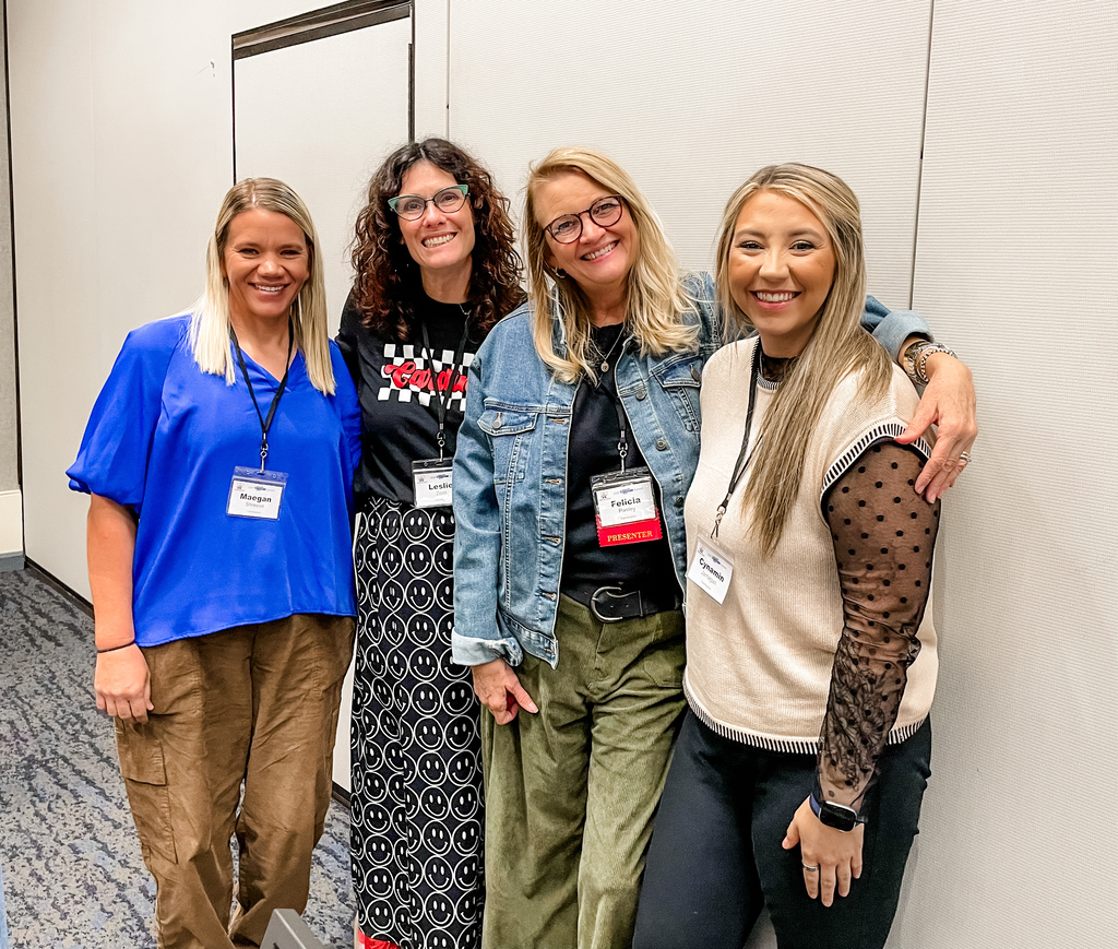 Four women in a room, two wearing glasses, all smiling and standing close together.
